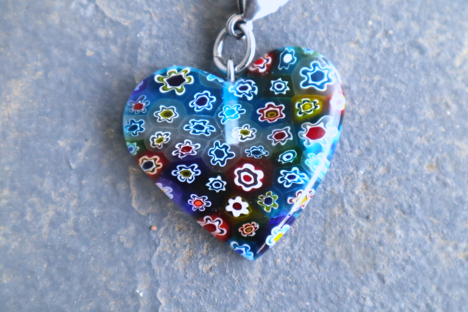 A heart-shaped multi-colored pendant with multi-colored flower patterns. A silver clasp is attached to the top of the pendant, resting on a gray surface.