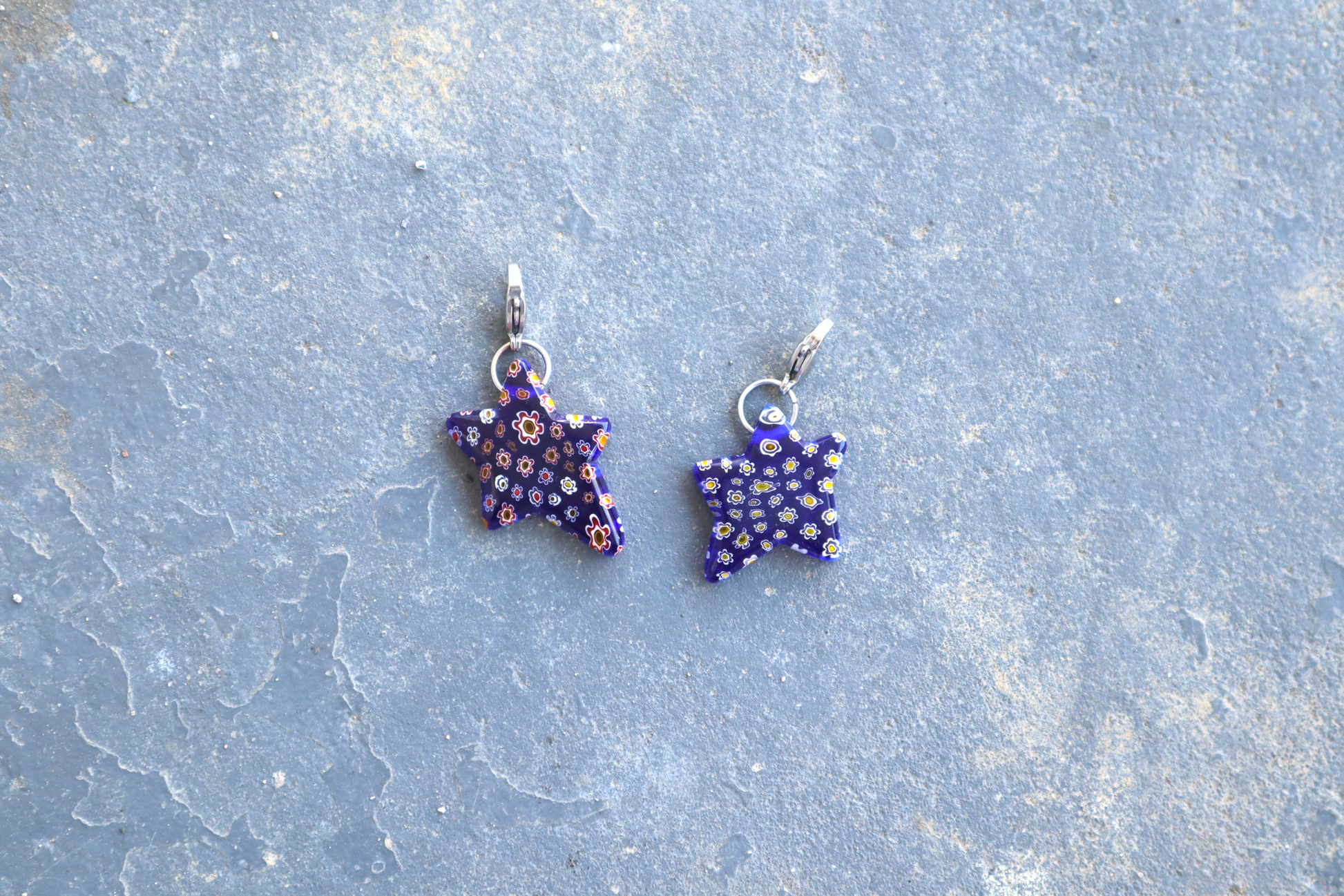 2 Star-shaped blue pendants with multi-colored flower patterns. A silver clasp is attached to the top of the pendant, resting on a gray surface.