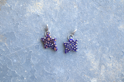 2 Star-shaped blue pendants with multi-colored flower patterns. A silver clasp is attached to the top of the pendant, resting on a gray surface.