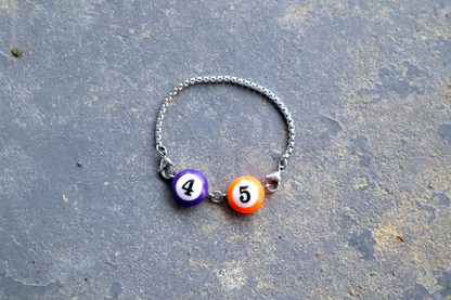 Silver chain bracelet with a purple, and orange billiard ball pendant marked "4", and "5" laid against a blue/gray surface.