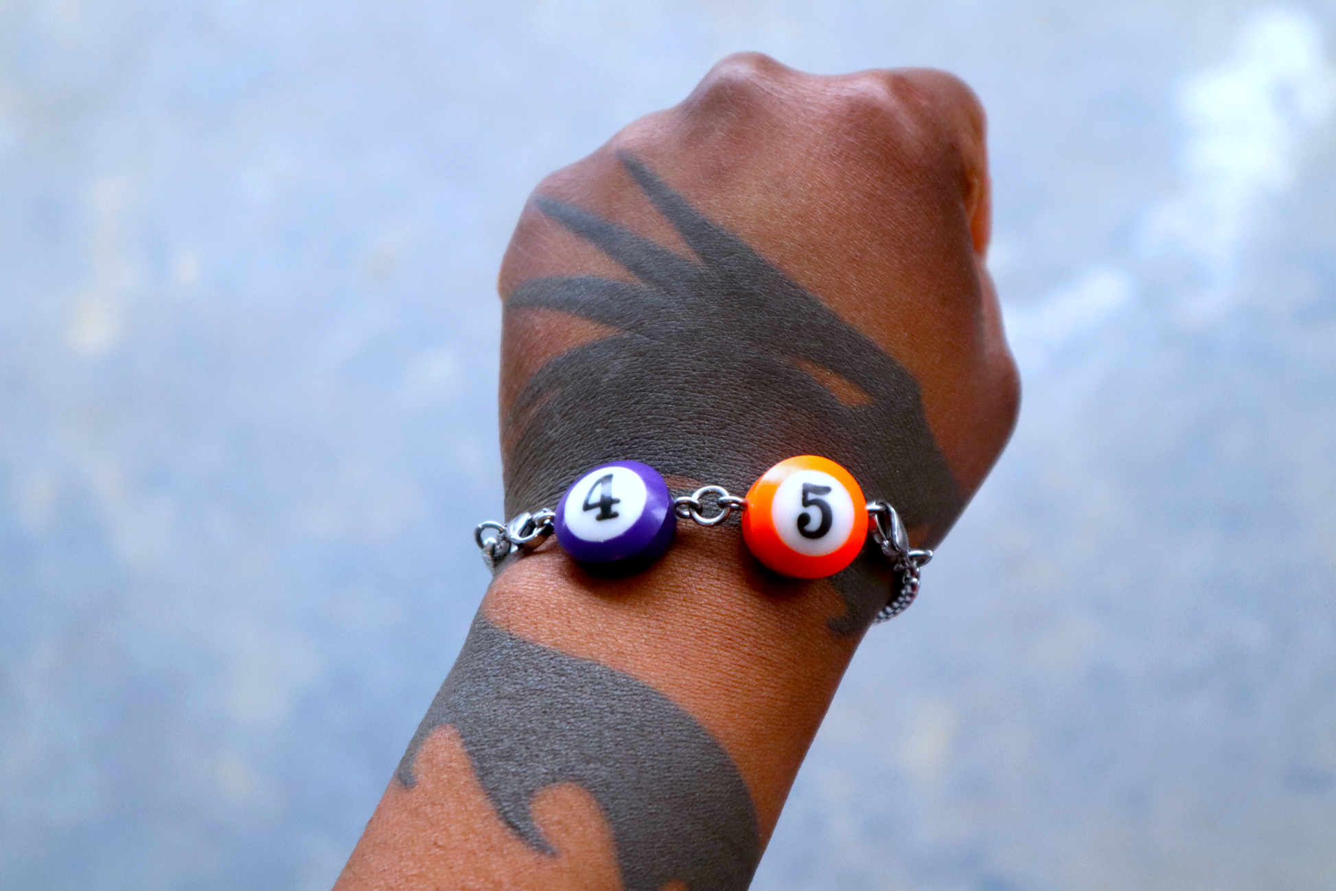 A hand with a dragon tattoo wears a bracelet of mini pool ball charms numbered 4, and 5.