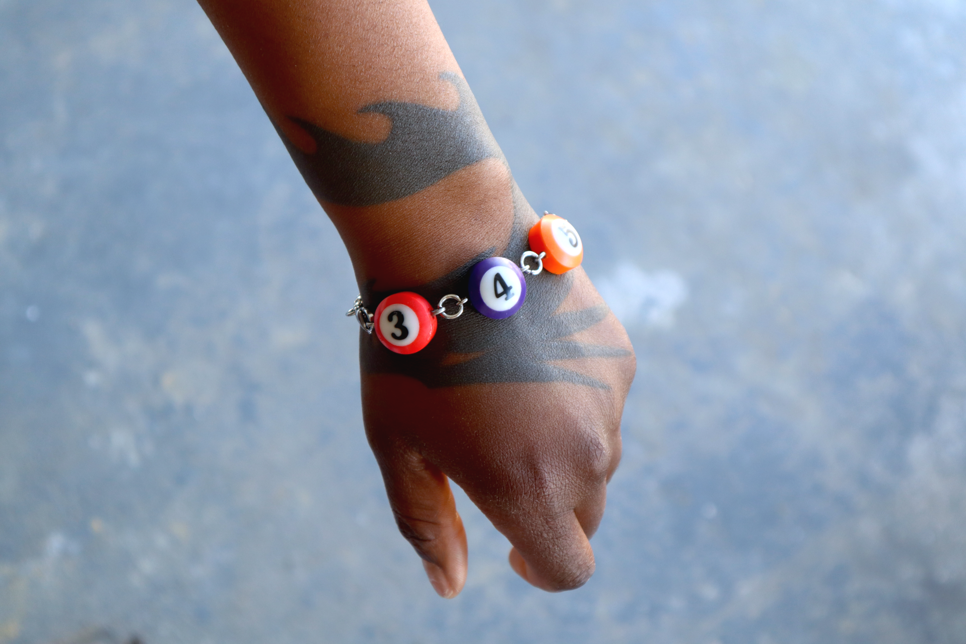 A hand with a dragon tattoo wears a bracelet of mini pool ball charms numbered 3, 4, and 5.
