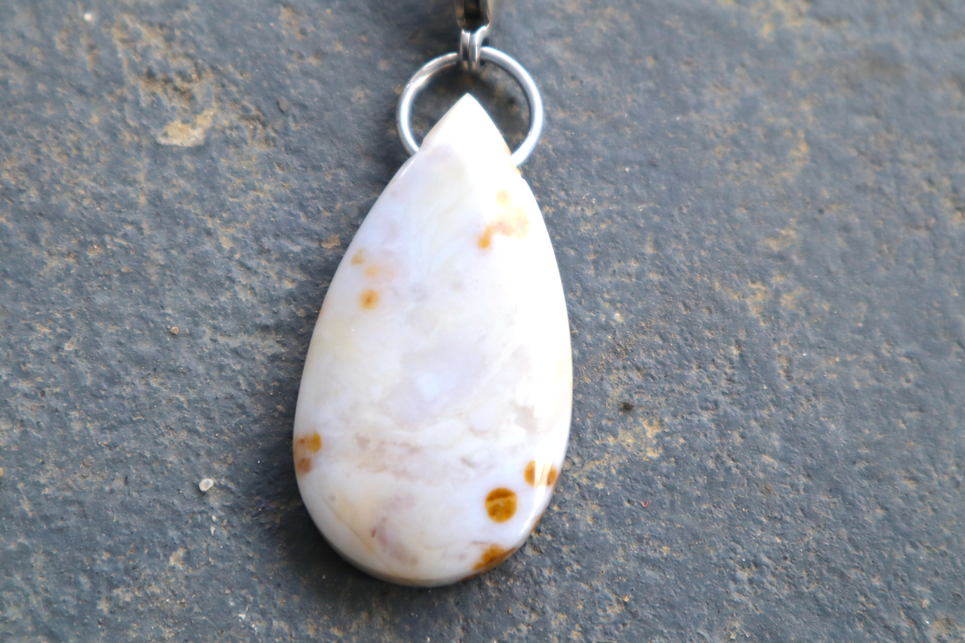  a close-up picture of a pointed oval-shaped stone and a tear drop-shaped stone, with a stainless-steel clasp attached to it, against a blue/grey stone background. The stone pendant is light grey, light blue, and white in color, with brown dots scattered across its surface. 