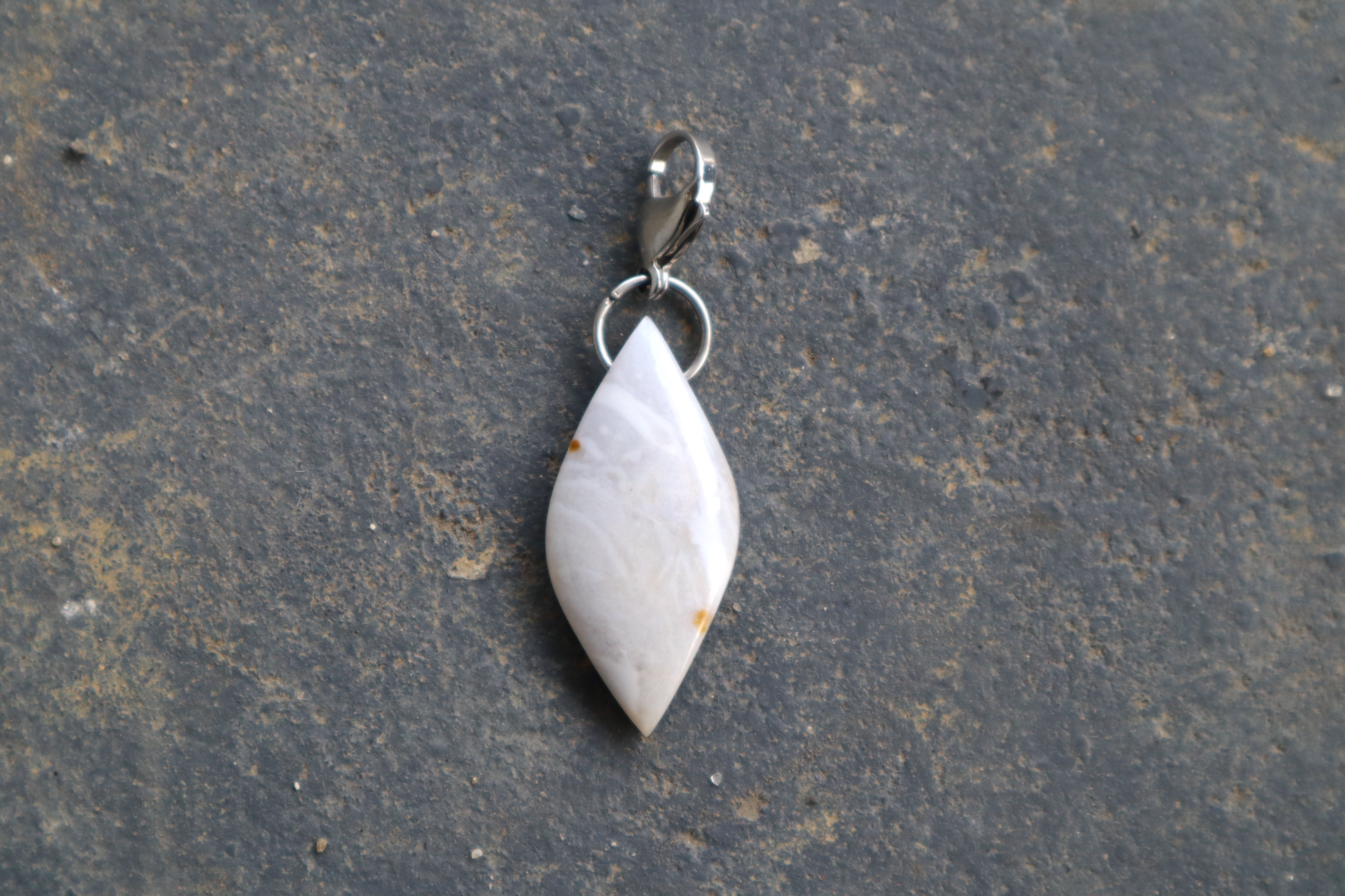 a pointed oval-shaped stone with a stainless-steel clasp attached to it, against a blue/grey stone background. The stone pendant is light grey, light blue, and white in color, with 2 brown dots on its surface. 