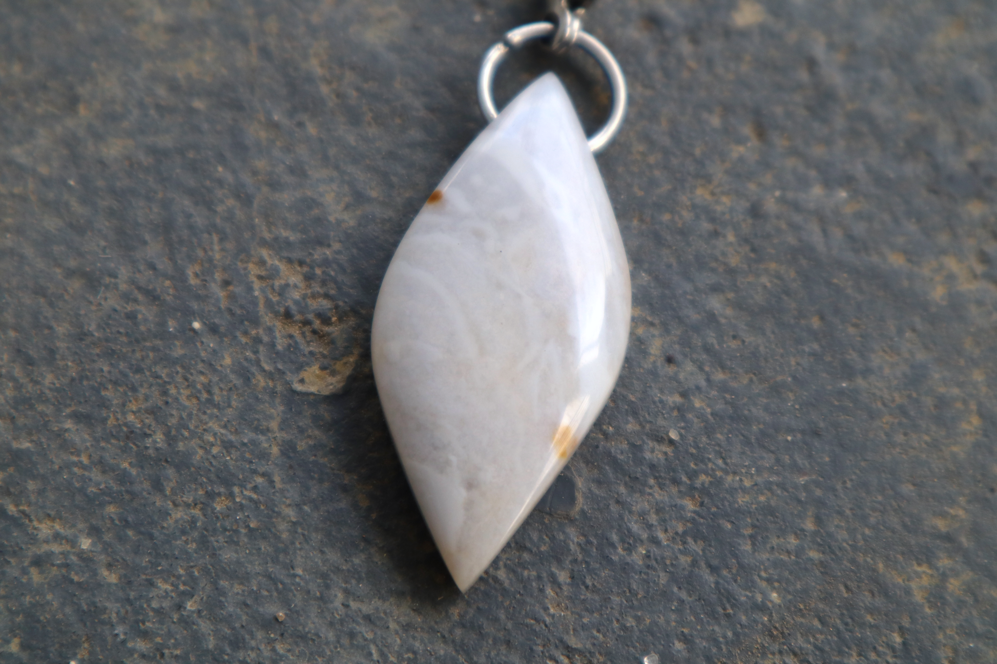  a close-up picture of a pointed oval-shaped stone, with a stainless-steel clasp attached to it, against a blue/grey stone background. The stone pendant is light grey, light blue, and white in color, with 2 brown dots on its surface. 