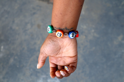 A hand wears a bracelet of mini pool ball charms numbered 14, 13, 2, and 3. The bracelet has these charms wrapped around the entire wrist.